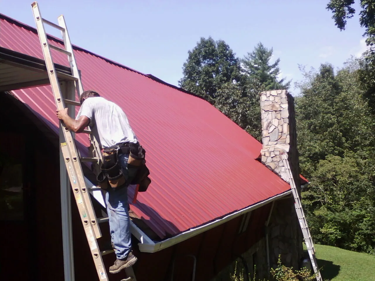 Roofer on a ladder beside a red metal roof and stone chimney — Soffit & Fascia Repair by Roofing Brothers in Baker