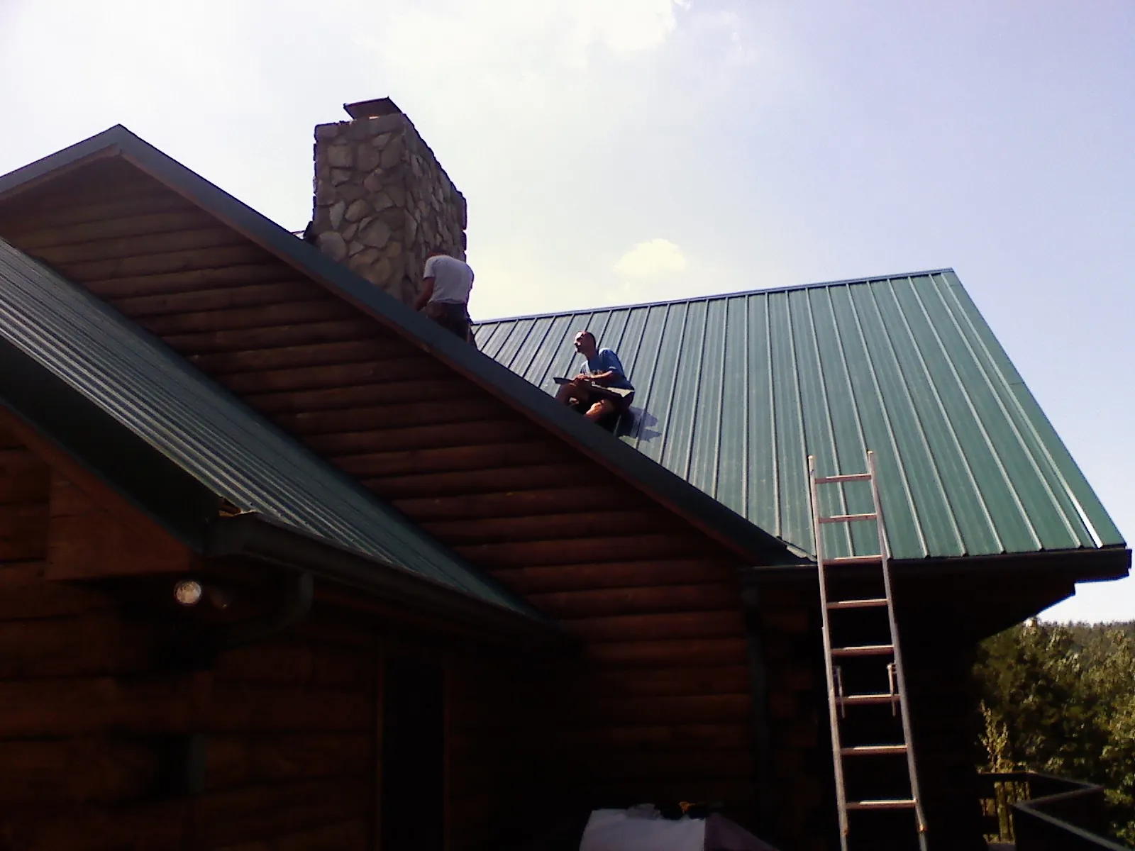Roofers installing green metal panels beside a stone chimney on a log home in Baker
